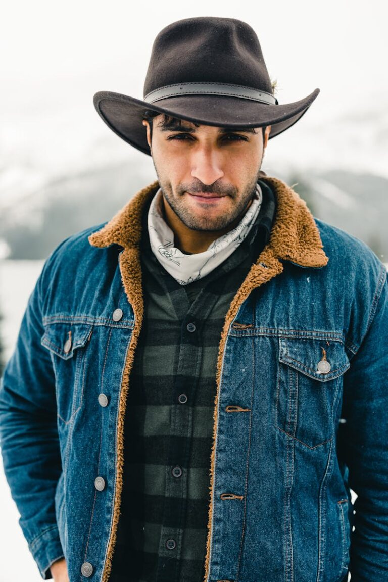 A rugged man in a denim jacket and cowboy hat stands against a snowy mountain backdrop in Jasper, Canada.
